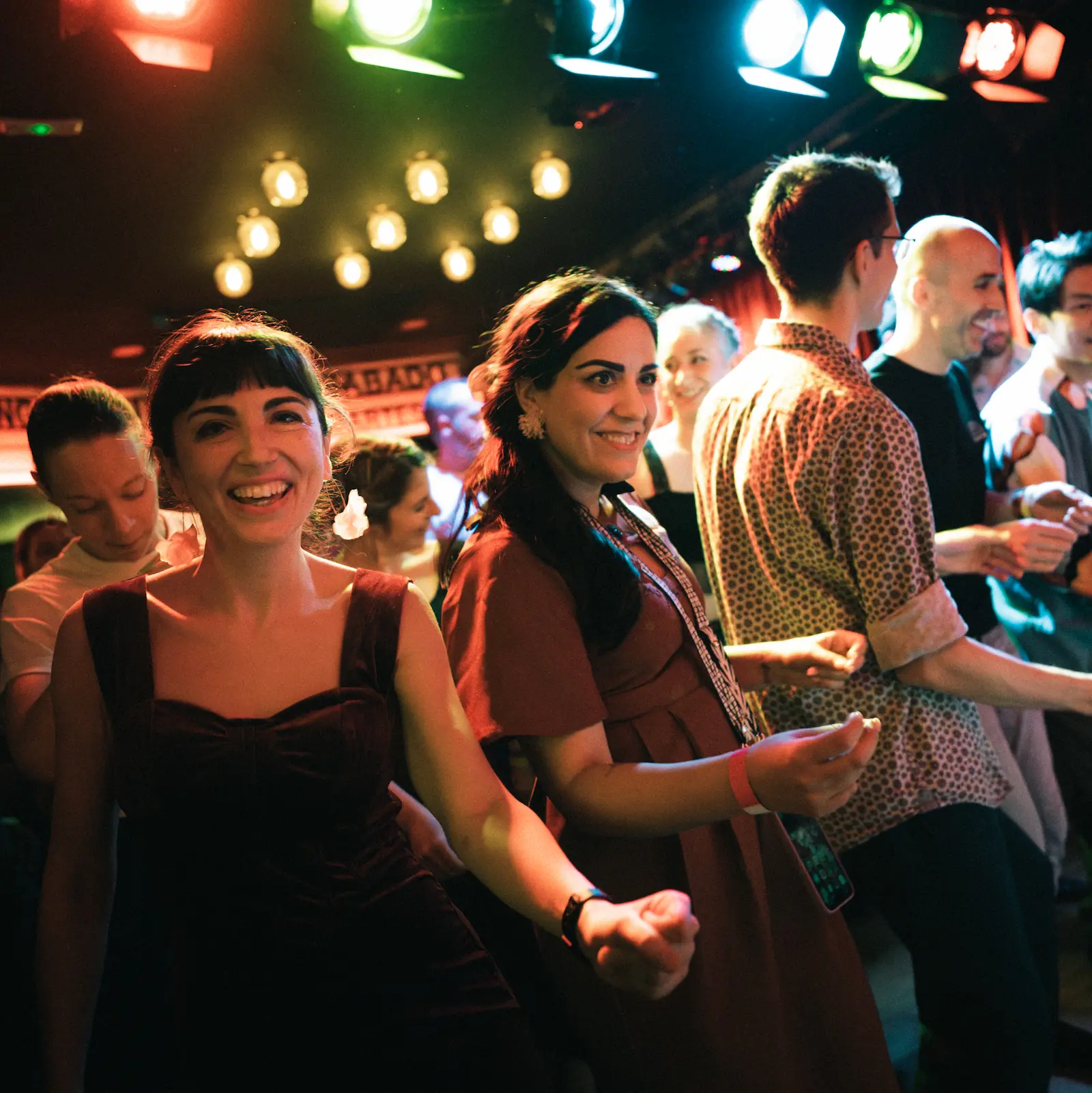 Dos mujeres sonríen y bailan entre una multitud bajo luces de colores en una sala de conciertos.