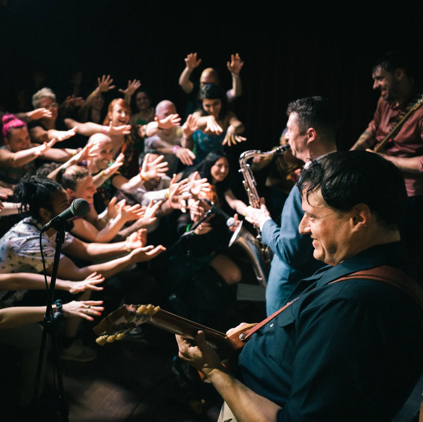 Músicos tocan guitarra y saxofón mientras el público extiende las manos hacia el escenario.